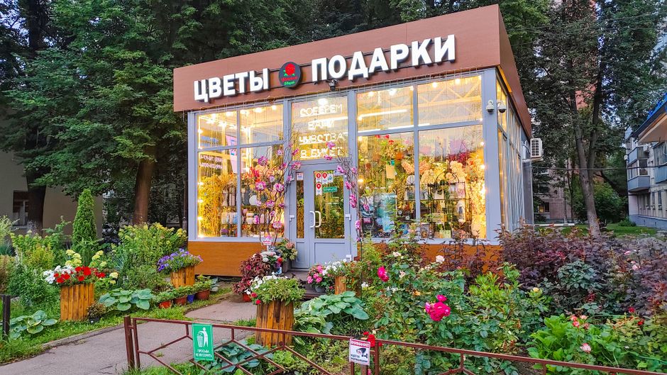 The exterior of a flower shop with large glass windows displaying vibrant floral arrangements inside. The shop has a light grey door and is framed by a variety of colorful potted plants and blooming f