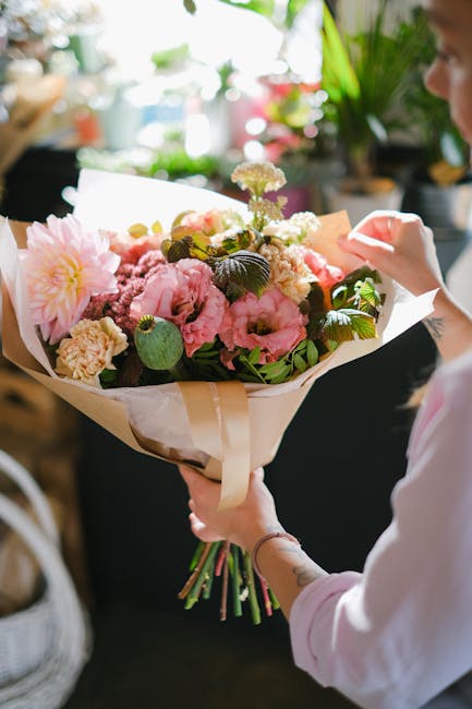 A person holding a large bouquet of fresh flowers including pink peonies, cream and white roses, and other blossoms, wrapped in white paper and tied with a beige ribbon. The arrangement features lush 