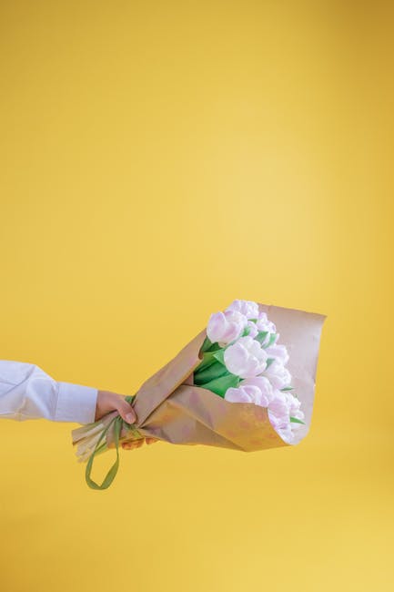 A hand holding a bouquet of fresh white tulips with soft pink accents, wrapped in light brown paper and secured with a green ribbon, set against a vibrant yellow background. The tulips are arranged in