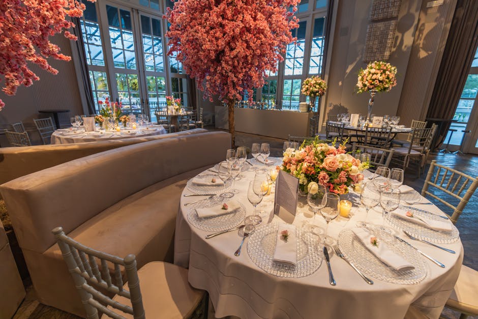 Elegant wedding reception tables decorated with floral arrangements featuring pink roses, white orchids, and greenery, set with clear glassware, white napkins, and silver cutlery. A large centerpiece 