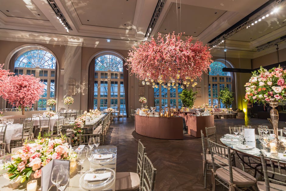An elegant wedding reception hall illuminated by soft ambient lighting, featuring large arched windows that allow natural light to filter in. The room is decorated with lush floral arrangements in sha